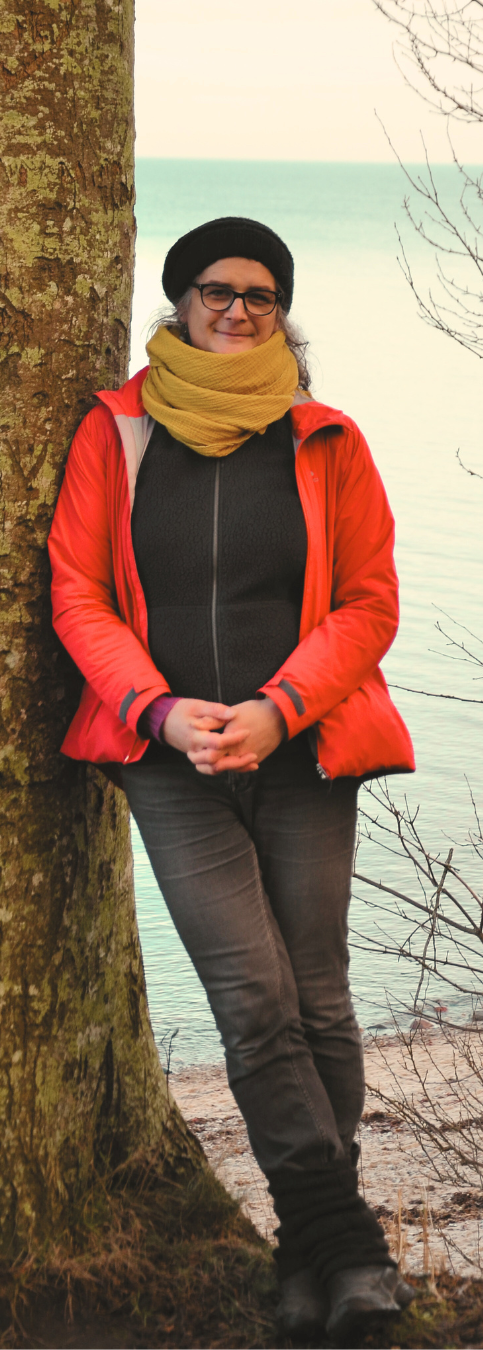 auf diesem bild bin ich als coachin zu sehen. ich stehe an einem baum gelehnt am meer. ich trage dunkle kleidung, eine leuchtendrote jacke.