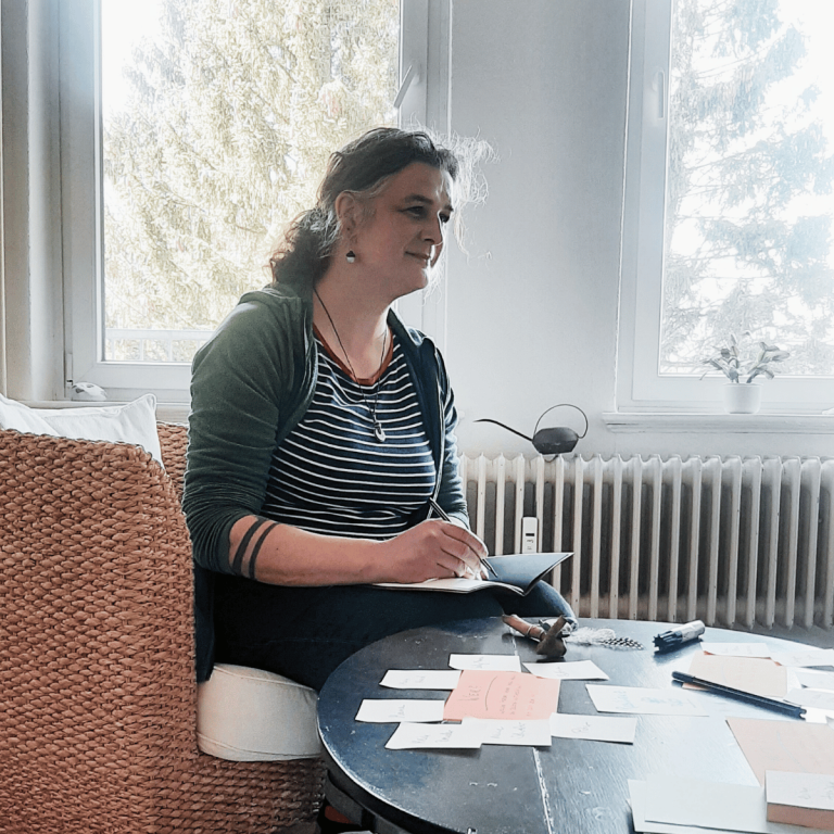 hierauf sieht man jana maria knoop, die auf einem sessel sitzt und einer person zuhört. auf dem tisch liegen einige beschriebene zettel.